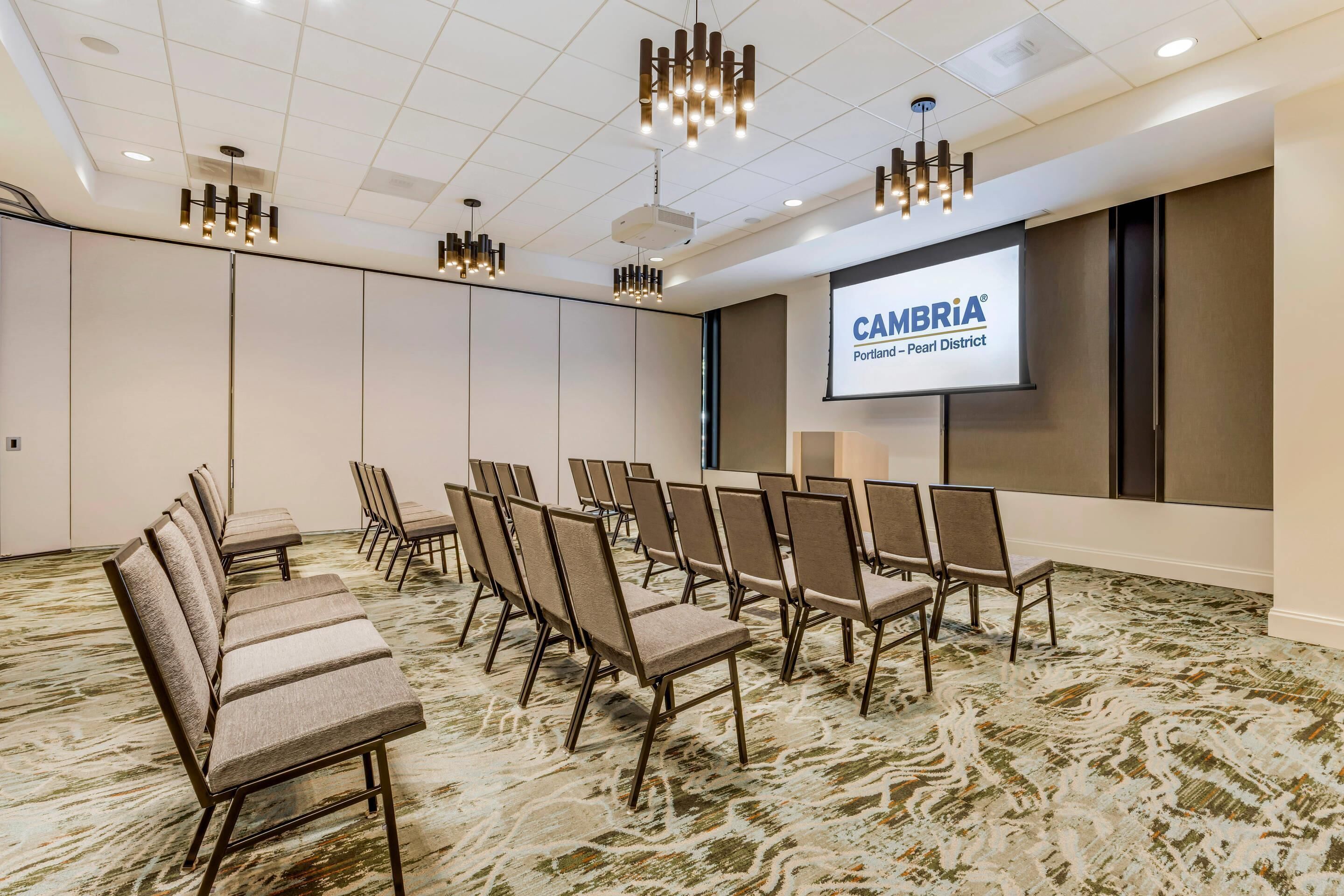 Ett konferensrum med rader av stolar, en projektorduk som visar bilden ”CAMBRIA Portland – Pearl District” och modern belysning.