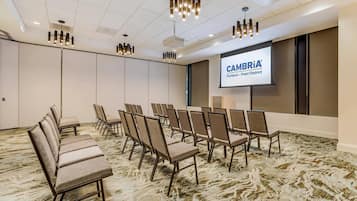 Ett konferensrum med rader av stolar, en projektorduk som visar bilden ”CAMBRIA Portland – Pearl District” och modern belysning.