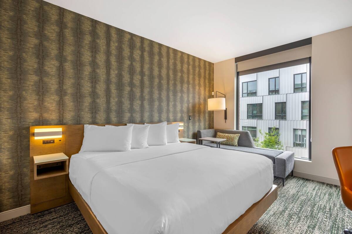 Une chambre d’hôtel avec un grand lit, une table de chevet avec une lampe, un petit canapé et une grande fenêtre donnant sur le bâtiment situé à l’extérieur.