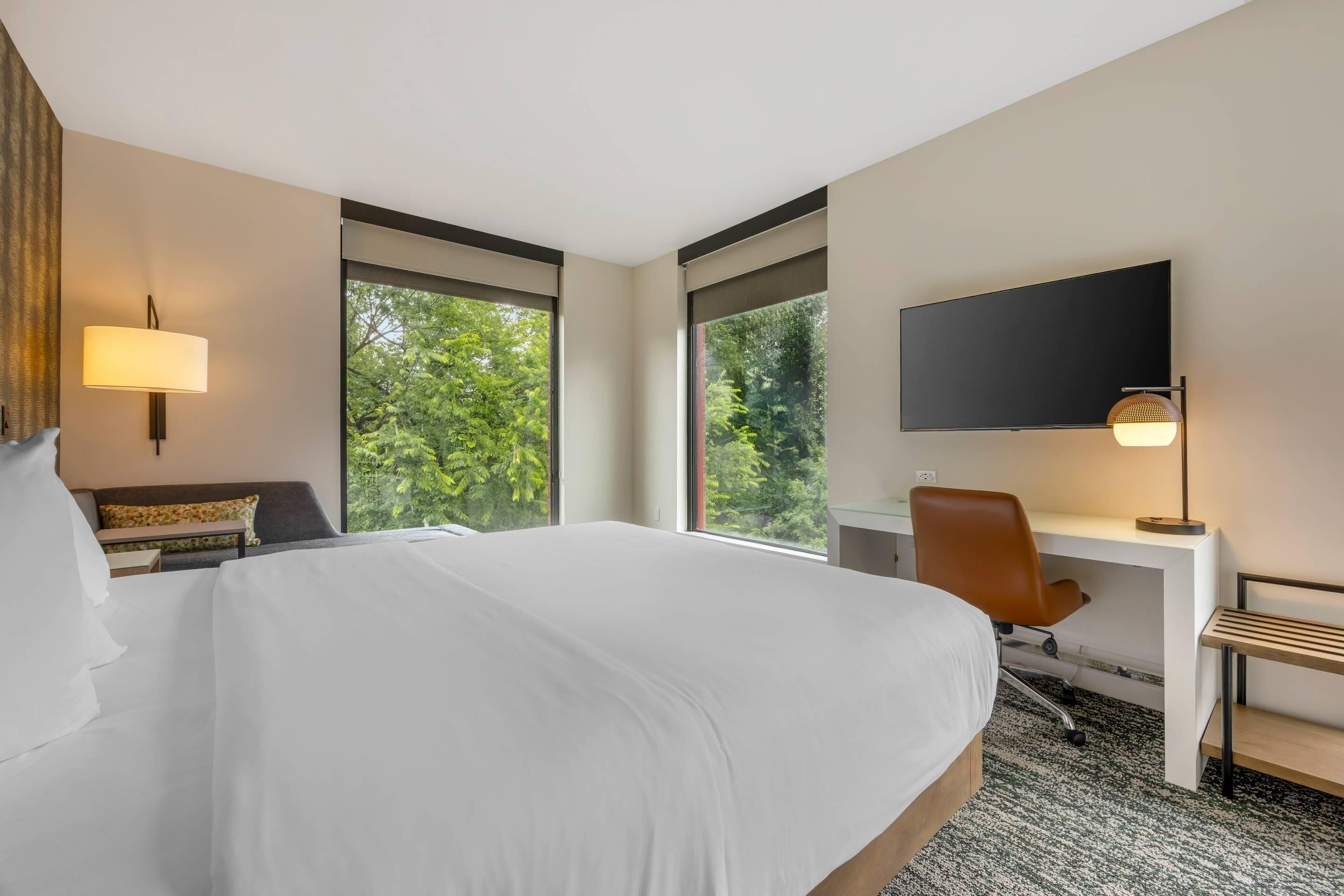 Une chambre d’hôtel avec un grand lit, un bureau, une chaise, une télévision et de grandes fenêtres donnant sur la verdure.