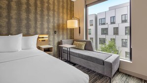 Une chambre d’hôtel avec un grand lit, un canapé, une petite table, une lampe et une vue sur un immeuble moderne, visible à travers une grande fenêtre.