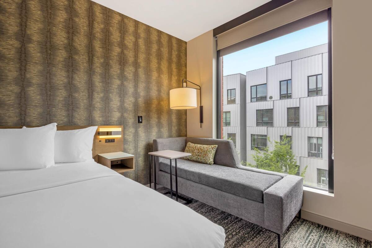 Une chambre d’hôtel avec un grand lit, un canapé, une petite table, une lampe et une vue sur un immeuble moderne, visible à travers une grande fenêtre.