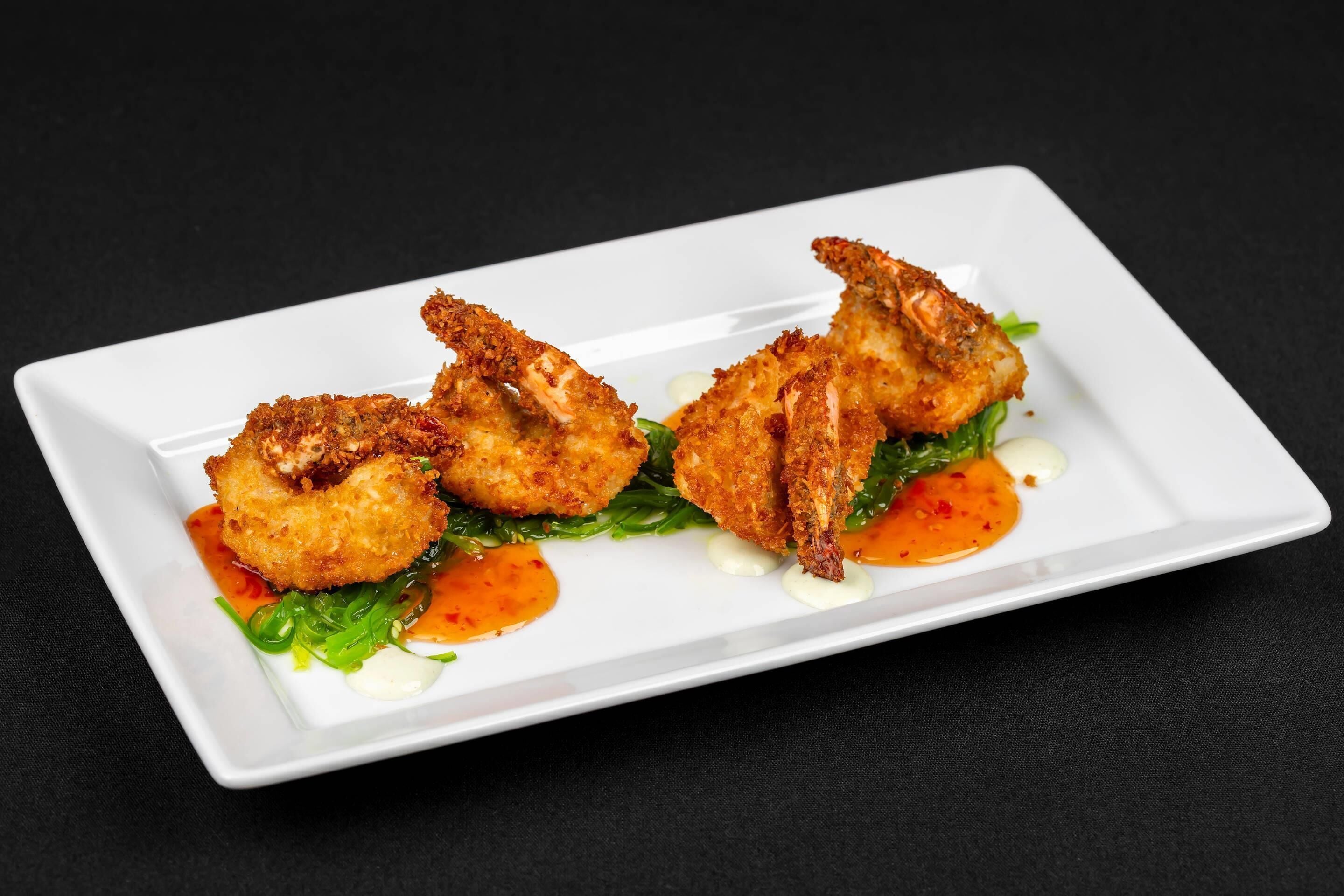 A white rectangular plate with three breaded shrimp on a bed of greens, accompanied by two dollops of sauce.