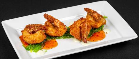 A white rectangular plate with three breaded shrimp on a bed of greens, accompanied by two dollops of sauce.