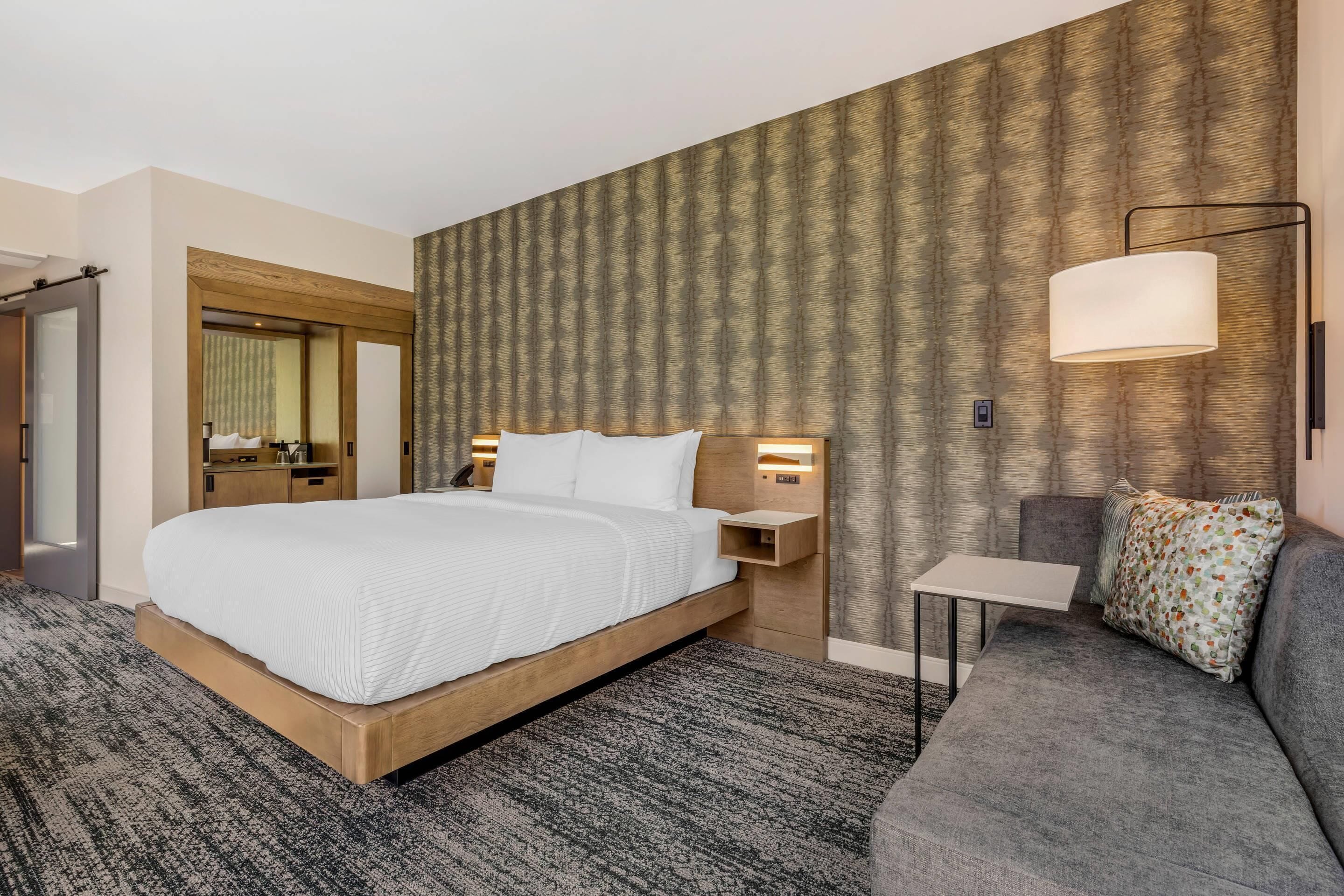 A hotel room with a large bed, a sofa, a small table, a wall-mounted lamp, and a mirror.