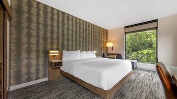 Habitación de hotel con una cama grande, una mesita de noche, un escritorio y una ventana con vistas a zonas verdes.