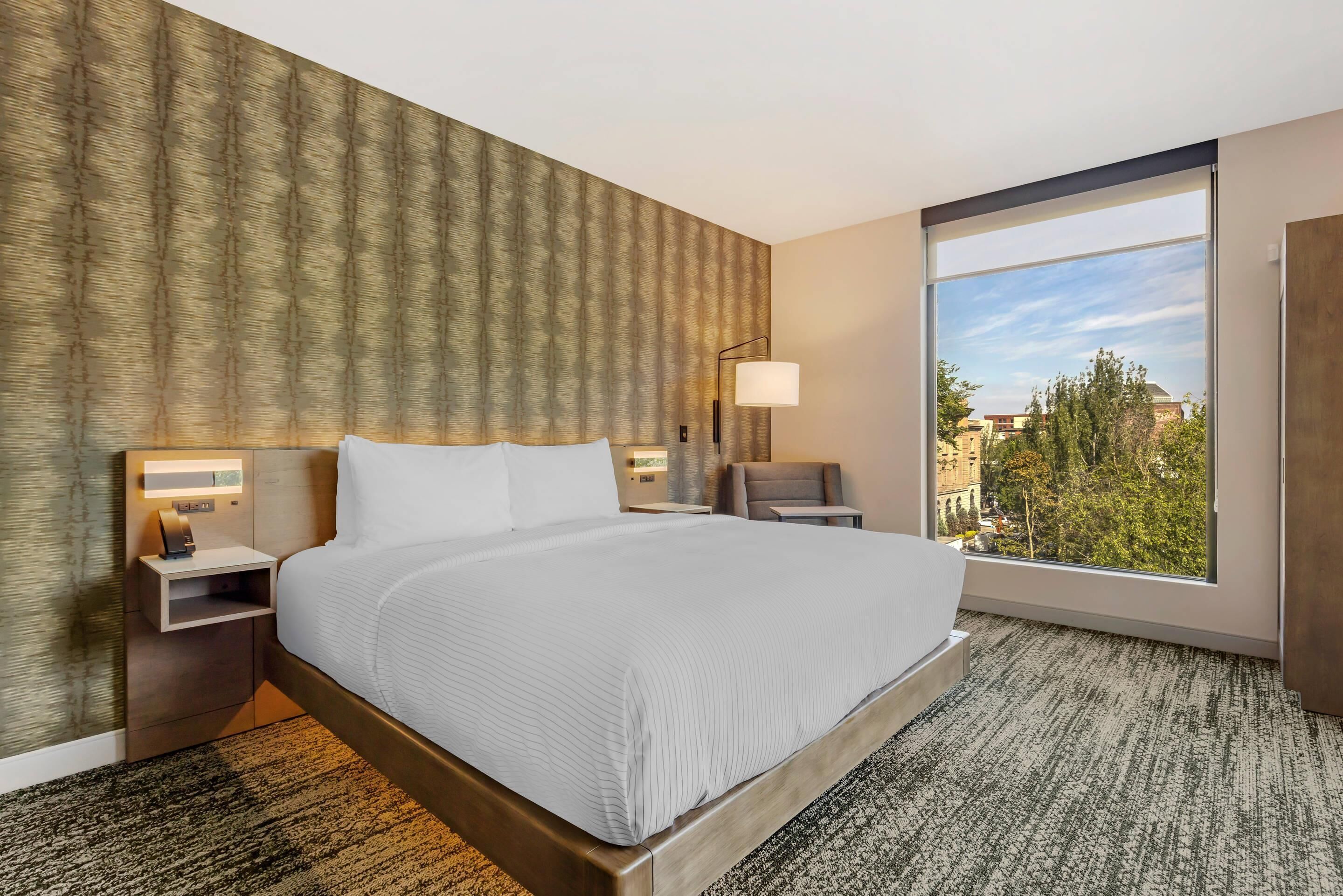 A hotel room with a large bed, a nightstand with a lamp, a view of the city through a large window, and a patterned carpet.