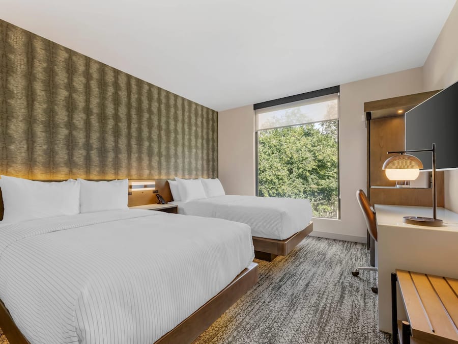 Habitación de hotel con dos camas, un escritorio, una silla, un ventanal grande y un papel tapiz estampado.