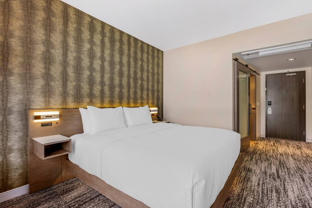 Une chambre d’hôtel avec un grand lit, une table de chevet avec une lampe, une tête de lit en bois et un sol recouvert de moquette.