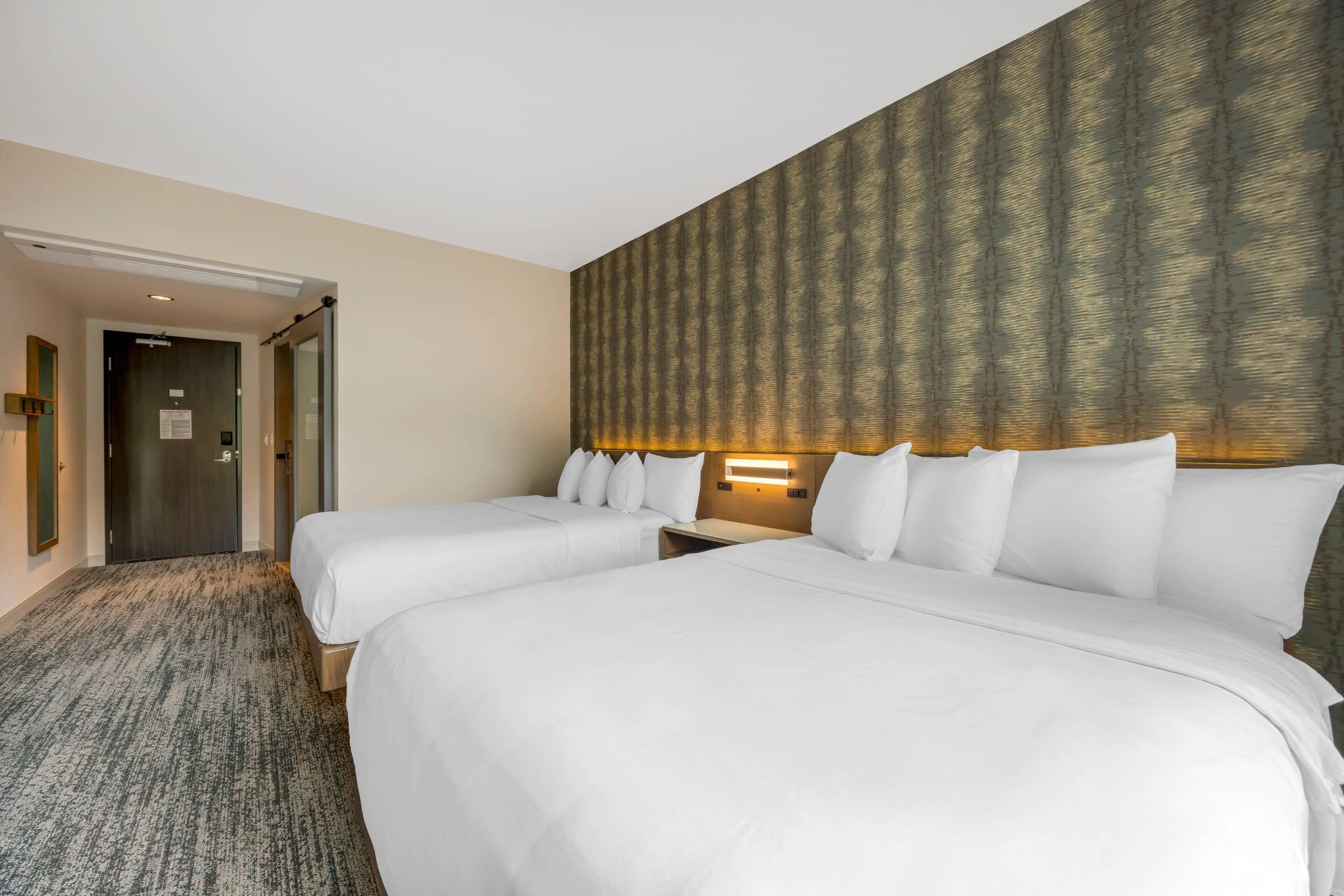 A hotel room with two beds, a carpeted floor, a wooden headboard with a patterned wallpaper, and a nightstand with a lamp.