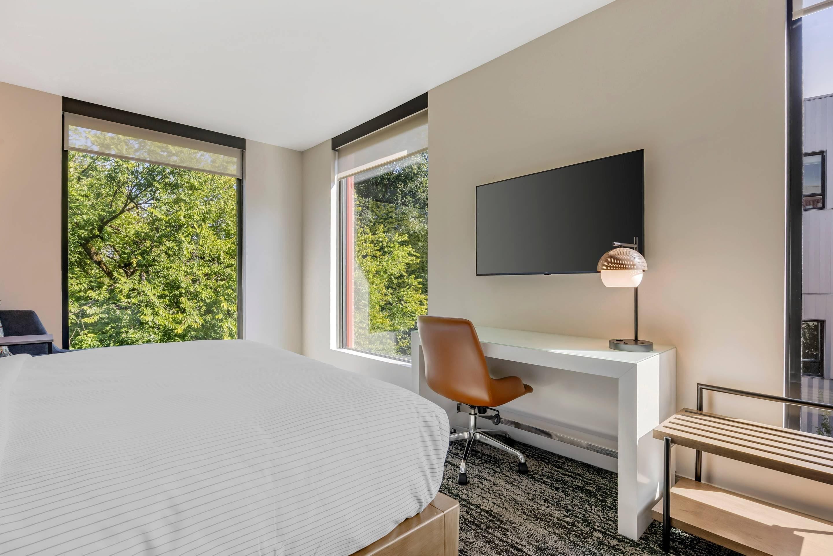 A modern hotel room with a large bed, a desk with a chair, a TV mounted on the wall, and large windows offering a view of trees.