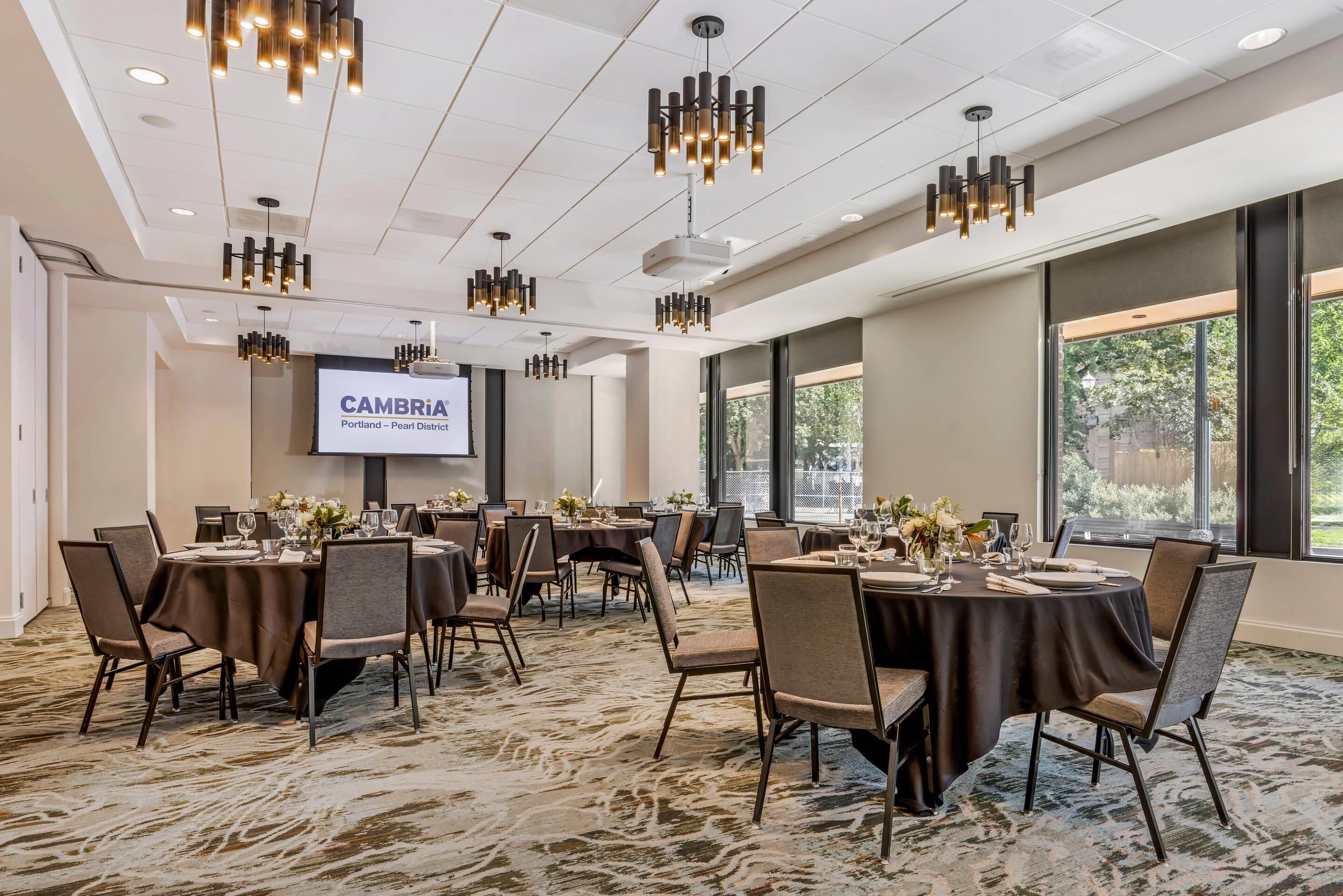 A conference room with multiple tables, chairs, and a projector screen displaying 'CAMBRIA Portland - Pearl District'.