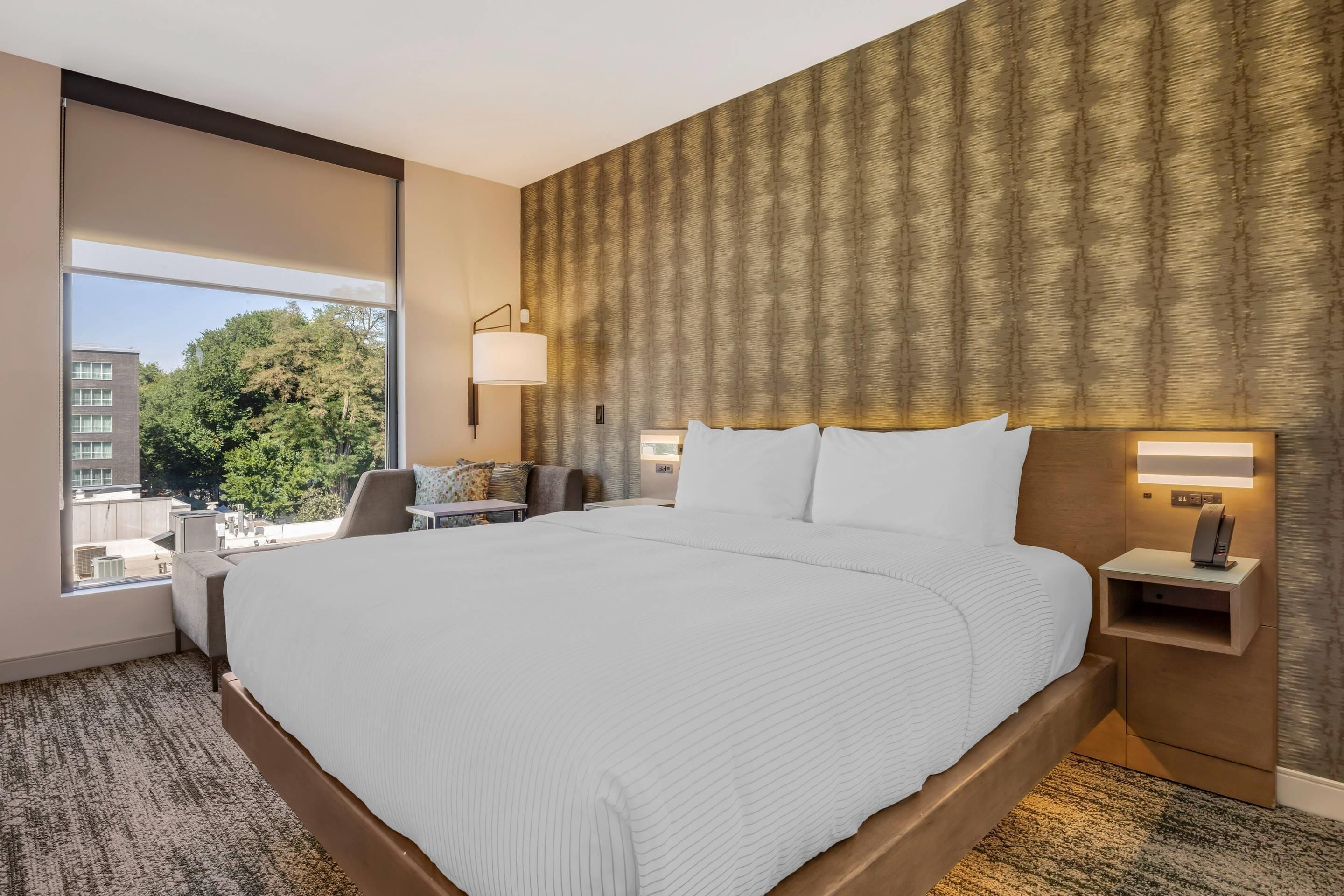 Habitación de hotel con una cama grande, una mesita de noche con lámpara, vistas a la ciudad y una ventana con vista a árboles.