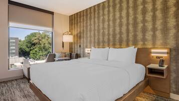 Habitación de hotel con una cama grande, una mesita de noche con lámpara, vistas a la ciudad y una ventana con vista a árboles.