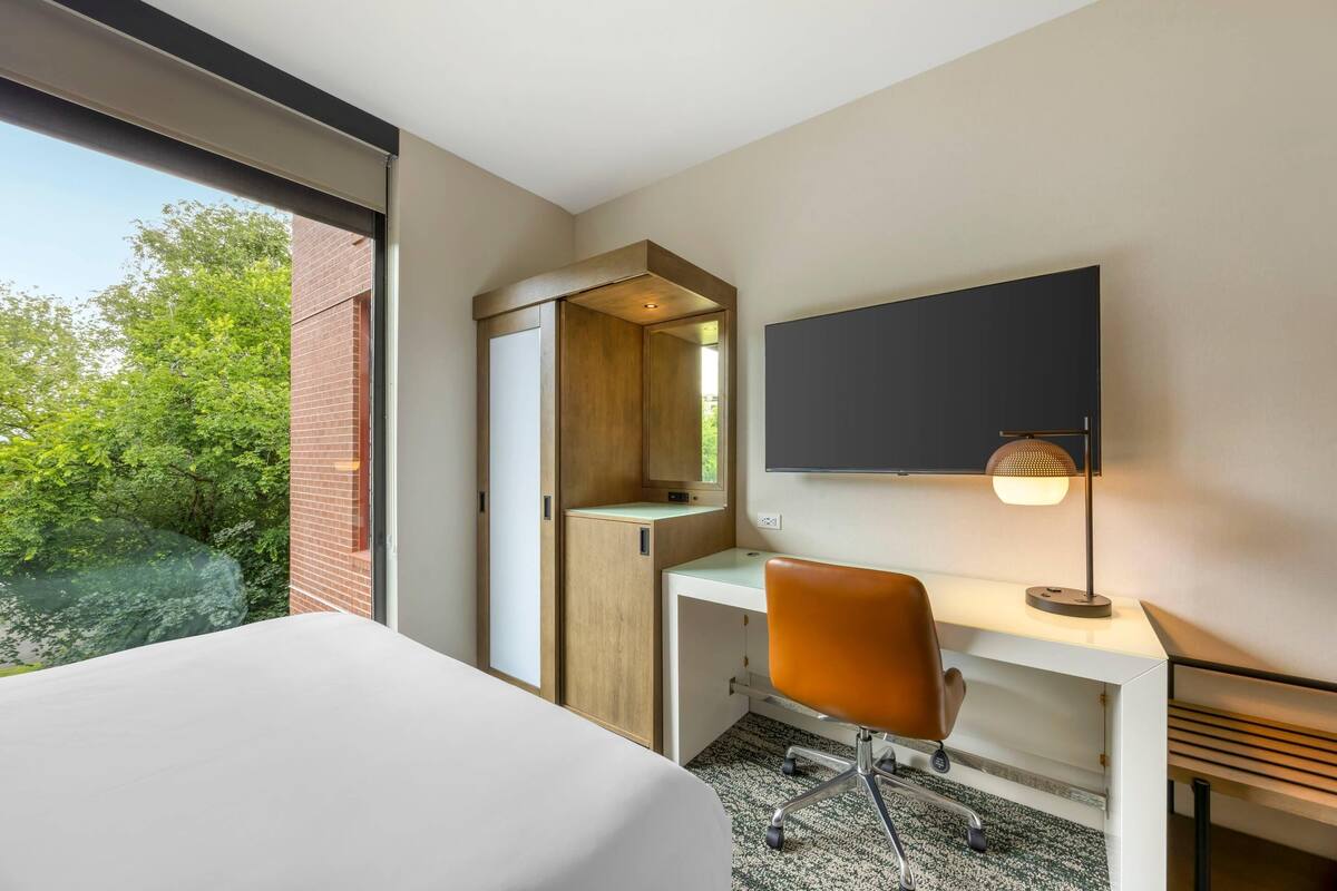 Une chambre d’hôtel comprenant un lit, un bureau avec une chaise, un téléviseur à écran plat et une fenêtre donnant sur un paysage verdoyant.