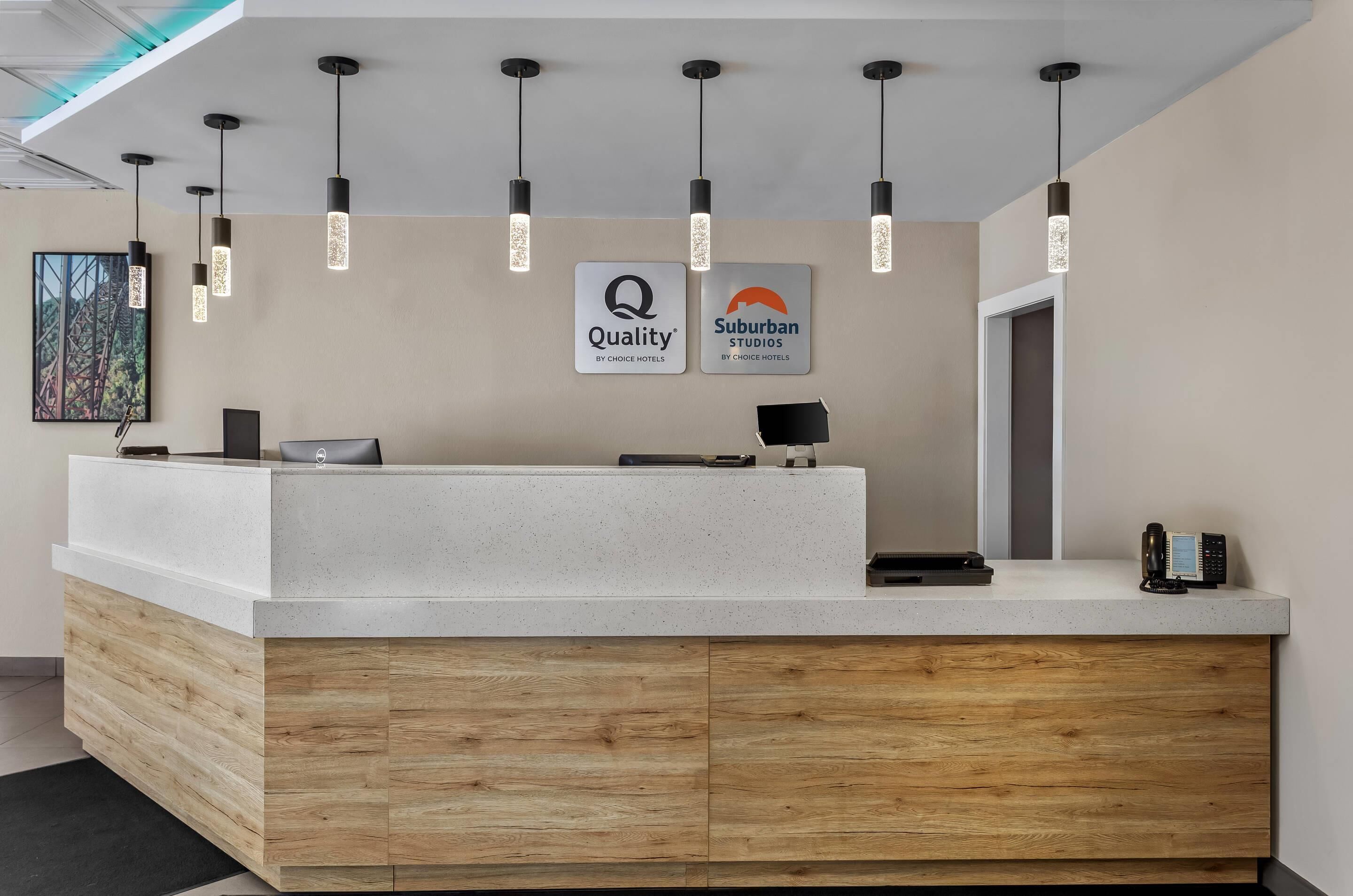 A reception desk with a modern design, featuring a white countertop and wooden base. There are two signs above the desk, one for 'Quality' and another for 'Suburban Studios', both by Choice Hotels.