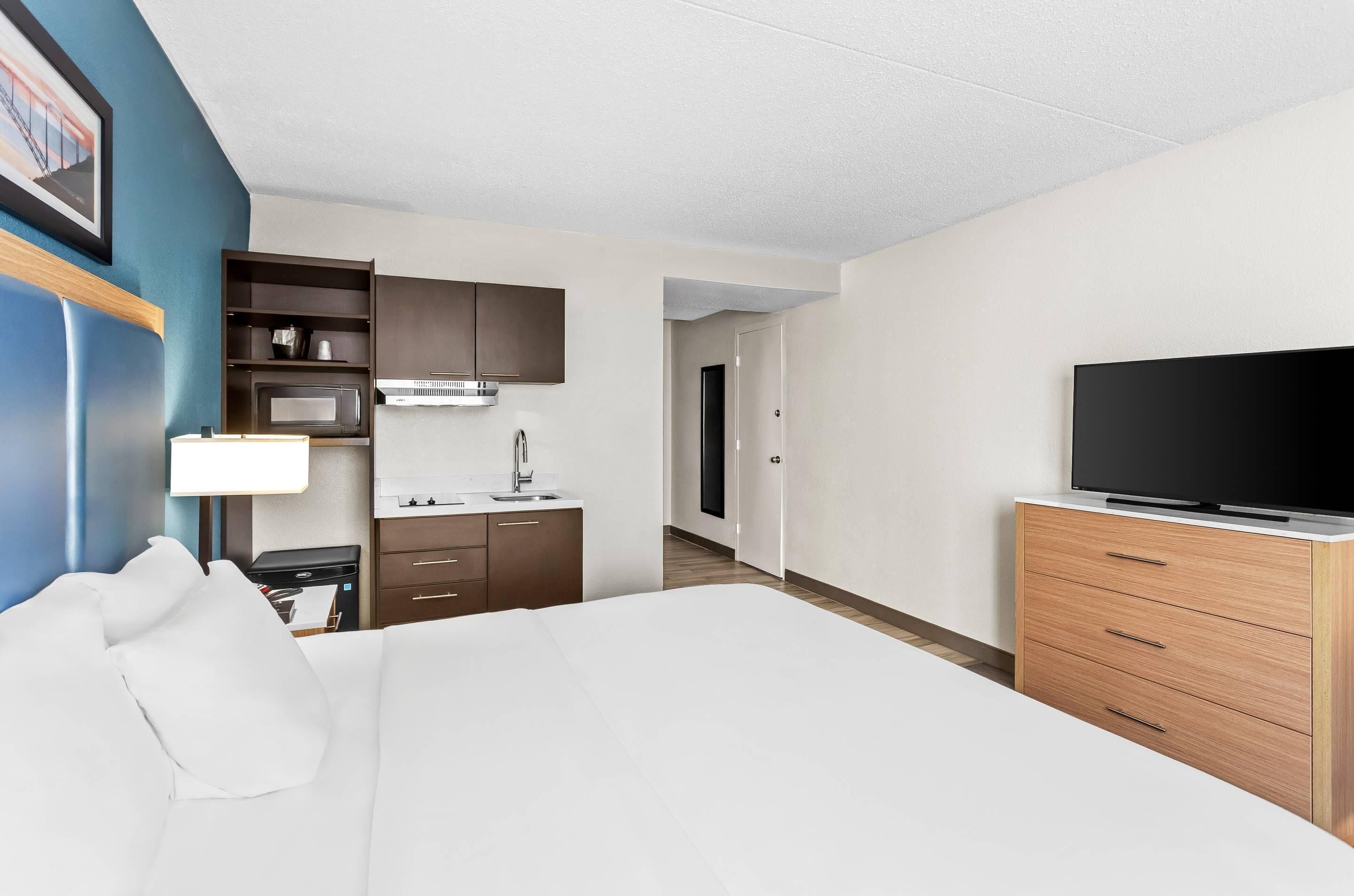 Habitación de hotel con cama, televisión, escritorio con lámpara, microondas y kitchenette pequeña.
