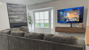 Un salon moderne avec un canapé gris, un téléviseur à écran plat fixé au mur et une fenêtre équipée de stores.