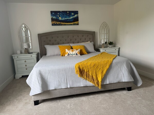 A bedroom with a large bed, two nightstands, a painting on the wall, and a window with a decorative frame.
