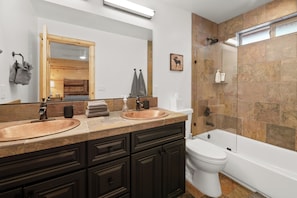 Un baño con dos lavamanos, muebles oscuros, un espejo grande, un inodoro y una ducha con mampara de cristal.