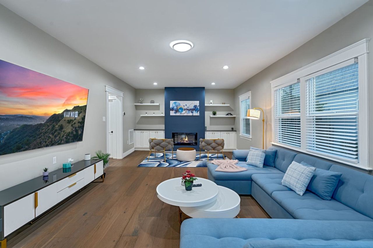 A modern living room with a fireplace, blue sofas, a flat-screen TV, and a large painting of a sunset.