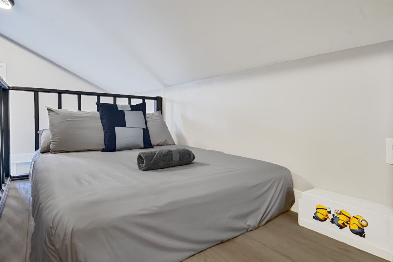 Una cama individual con cubrecolcha y almohadas grises, una toalla gris enrollada y un estante de pared con una decoración temática de Minions.