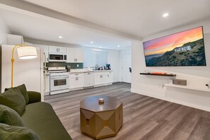 Una sala de estar moderna con un sofá, una mesa de centro redonda, un televisor de pantalla plana montado en la pared y una zona de cocina al fondo.
