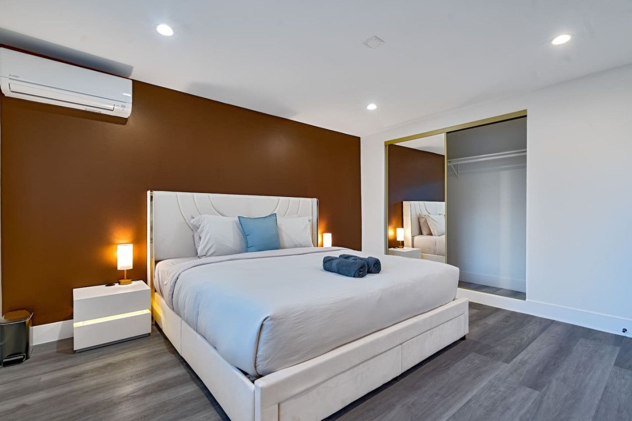 A modern bedroom with a large bed, bedside tables, an air conditioning unit, and a mirror.