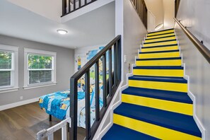 Un escalier aux marches jaunes et bleues, un lit recouvert d’une courtepointe bleue et jaune, et une fenêtre avec des stores.