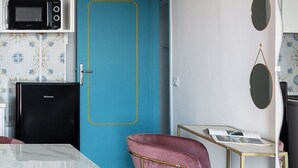 Une cuisine compacte équipée d’un four à micro-ondes, d’un réfrigérateur et d’une porte bleue. On y trouve également un coin repas avec une table blanche et des chaises roses.