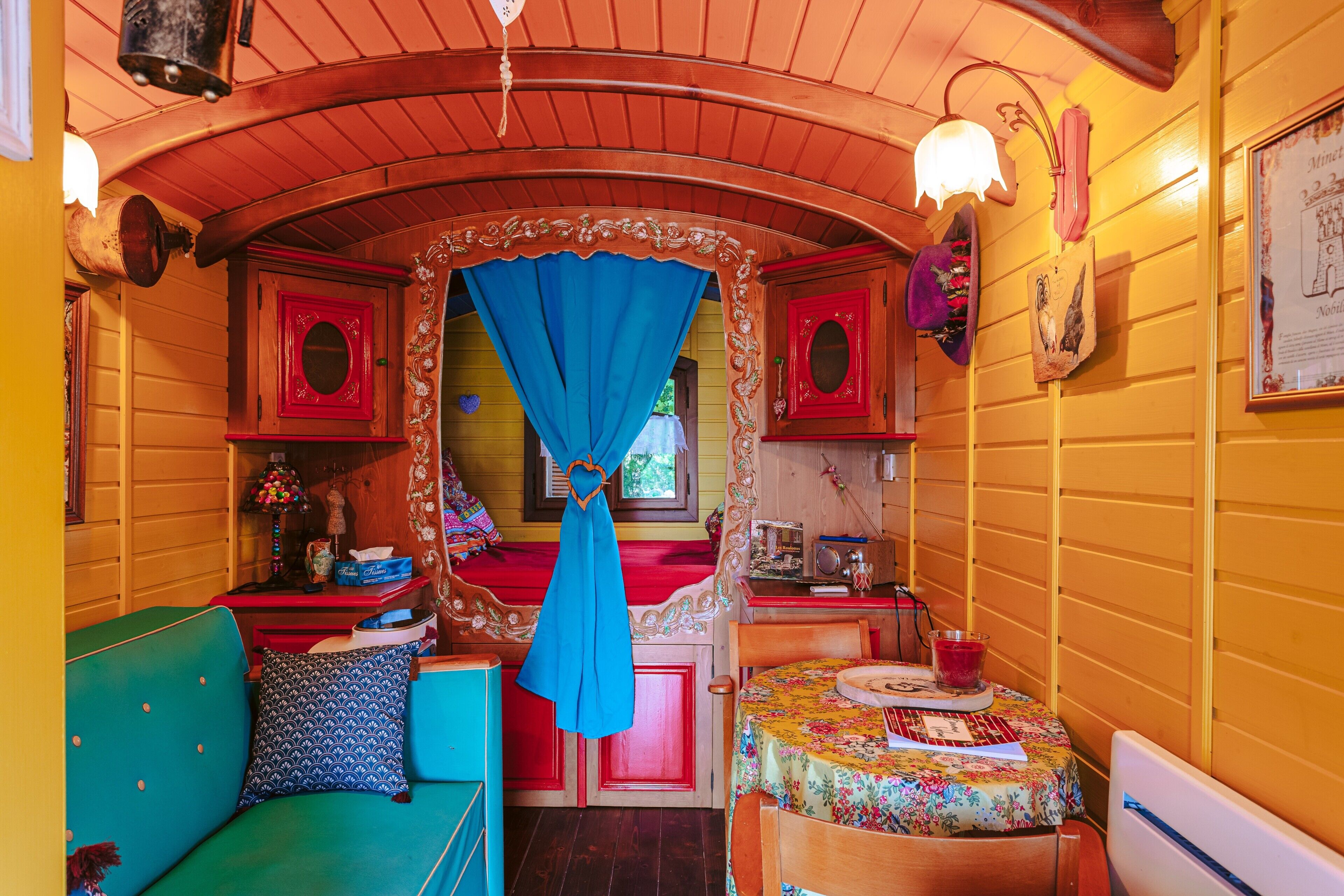 A cozy room with a curved ceiling, a bed with a blue canopy, a small table with a floral tablecloth, and a red cabinet.