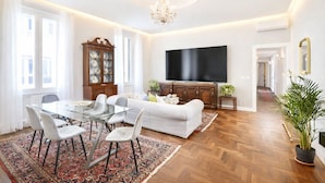 Un vaste salon comprenant une table à manger en verre, des canapés blancs, un grand téléviseur à écran plat, un meuble en bois et un lustre.