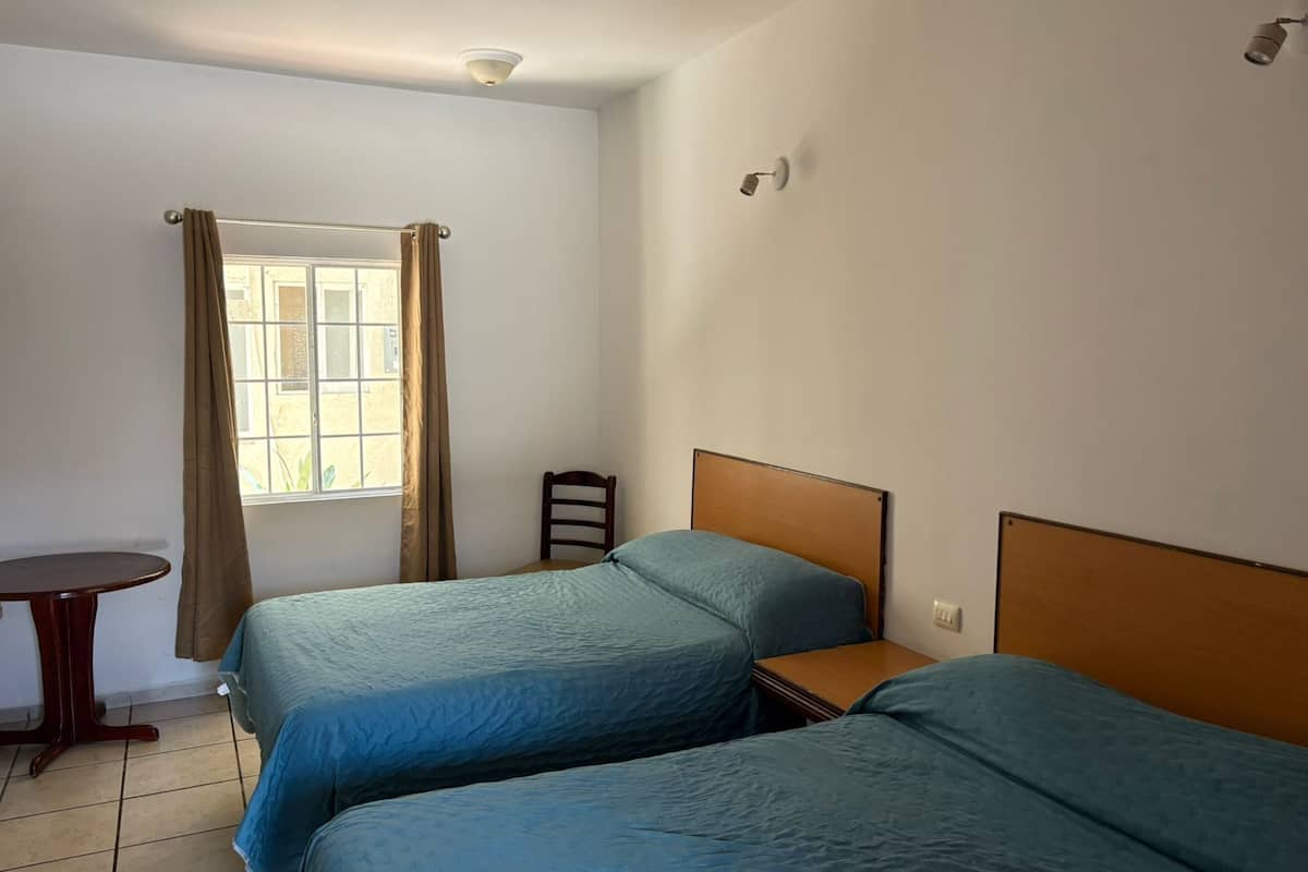 A room with two beds, a small table, a chair, and a window with curtains.