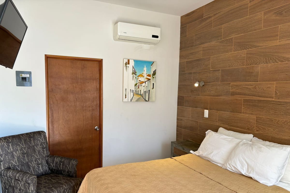 A bedroom with a wooden headboard, a bed with white linens, a patterned armchair, a door, and a wall-mounted air conditioner.