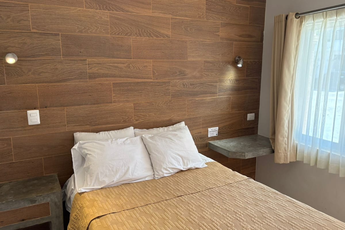 A bedroom with a wooden headboard, a bed with white bedding, a bedside table, and a window with curtains.