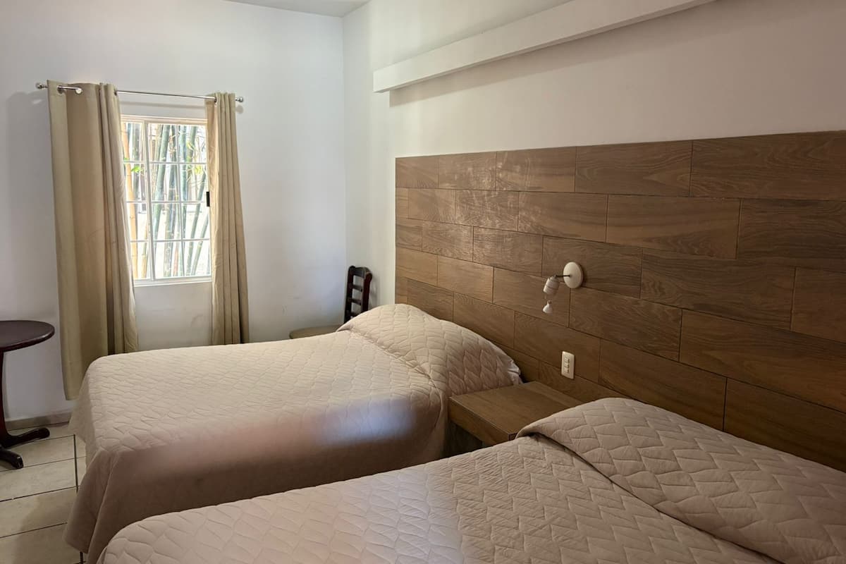 A hotel room with two beds, a wooden headboard, a small table, and a window with curtains.