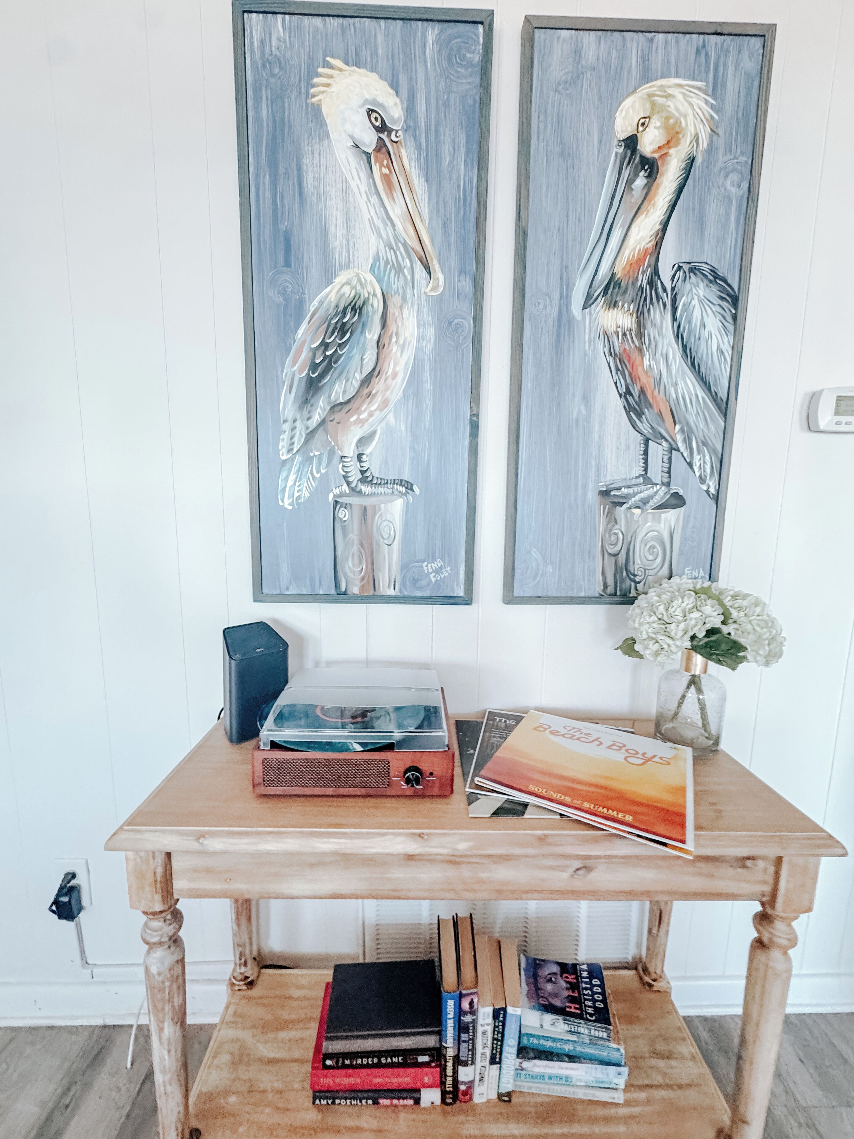 A wooden table with two paintings of pelicans, a vase with flowers, and various books.
