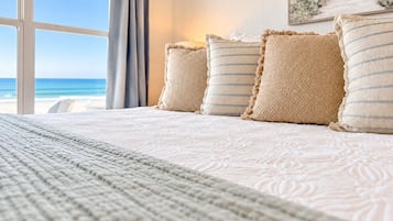 A bed with a patterned bedspread and pillows, a window with a view of the beach, and a painting on the wall.