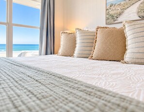 Un lit avec une couvre-lit à motifs et des oreillers, une fenêtre donnant sur la plage et un tableau accroché au mur.