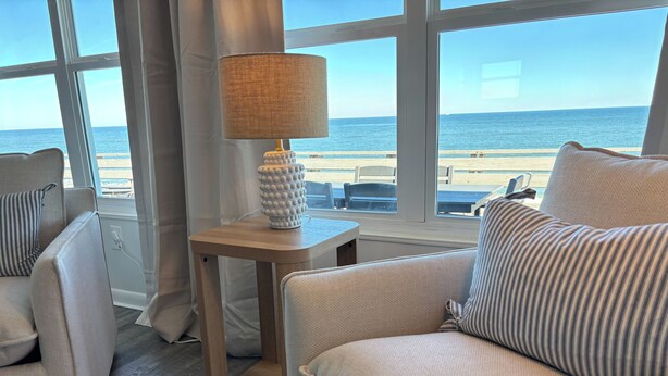Un coin salon confortable avec vue sur la plage grâce à de grandes fenêtres, une lampe posée sur une petite table en bois et un tapis à motifs sur le sol.