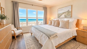 A bedroom with a large bed, a view of the beach, a wooden dresser, a chair, and a nightstand with a lamp.