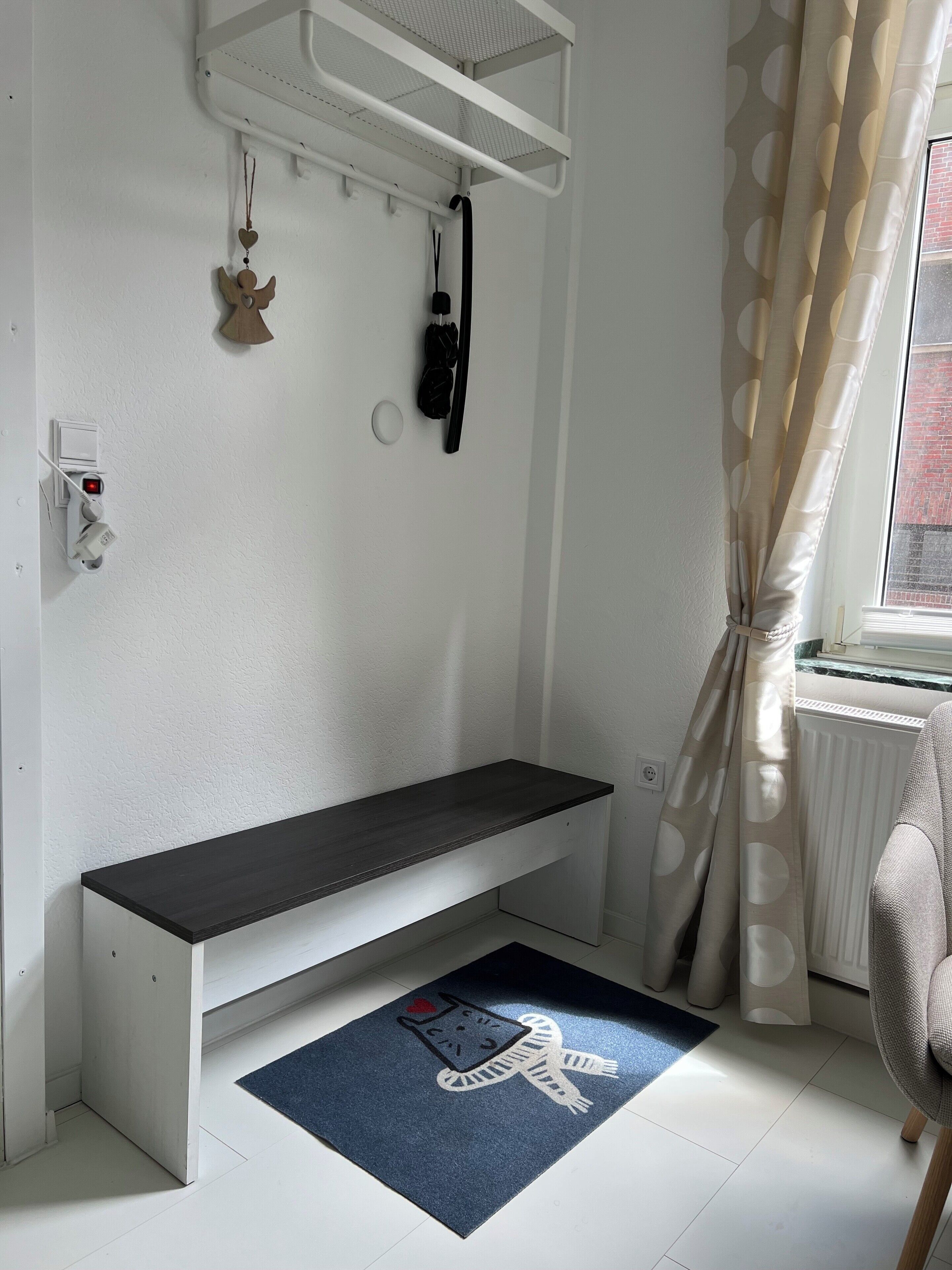 A room with a white wall, a black bench, a blue mat with a snowman design, a radiator, and a window with curtains.