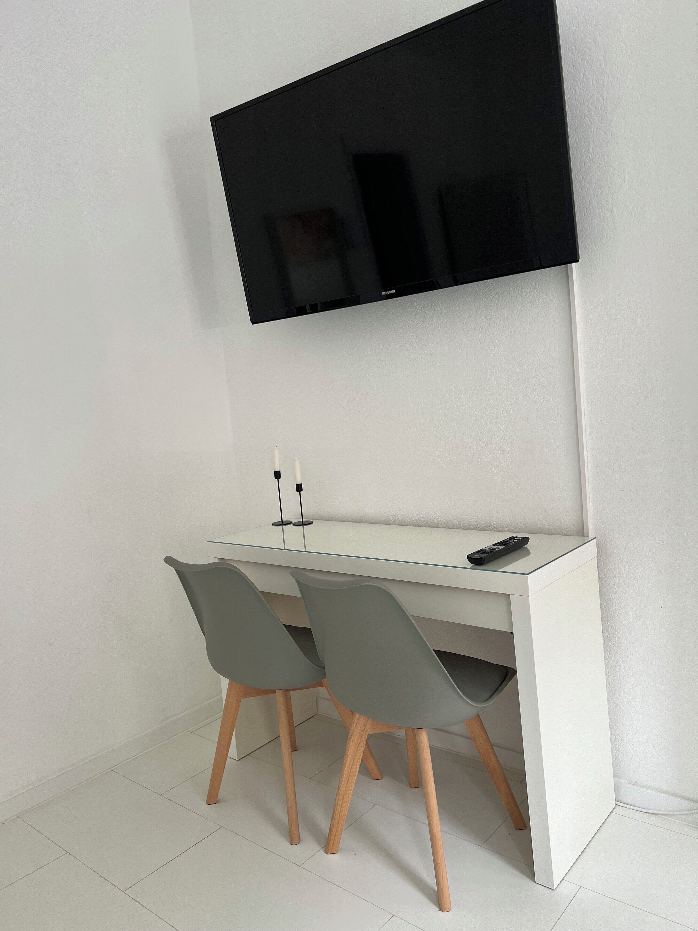 Un escritorio moderno blanco con dos sillas grises, un televisor de pantalla plana montado en la pared y un control remoto sobre el escritorio.