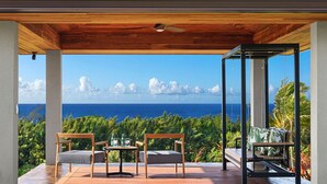 A covered outdoor seating area with a wooden deck, two chairs, a table, and a glass enclosure. - Kauai Oasis Luxe Ocean View, Pool SPA AC Sleep22 (Koloa)