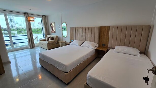 A hotel room with two beds, a sofa, a large window with a view of a body of water and buildings, and a hanging light fixture. - Loft Mr1-4g Facing the sea in a High Level Neighborhood (Cartagena)