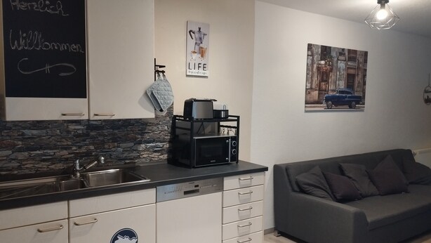 A modern kitchen with a stone backsplash, a blackboard with 'Herzlich Willkommen' written on it, a microwave, and a coffee machine on the counter. There's a gray sofa and a wall with a 'Life' poster and a vintage car picture. - Vacation apartment Hof Lanze with a view of the Eichenallee (Prezelle)