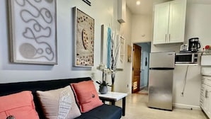 Una sala de estar moderna con un sofá negro, una mesita con un florero, y una cocina con armarios blancos y electrodomésticos de acero inoxidable.
