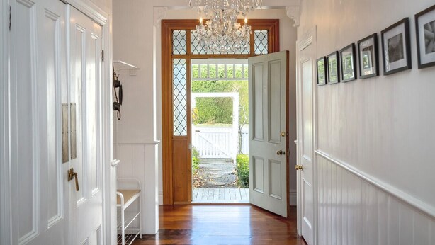 A hallway with white paneled walls, a wooden floor, and a chandelier. There are doors on the left and a door with a window at the end.