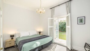 A bedroom with a bed, bedside lamps, a chandelier, a door leading to a garden, and a framed picture on the wall.