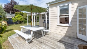 A wooden deck with a picnic table and umbrella, a white building with a window, and a fenced yard.
